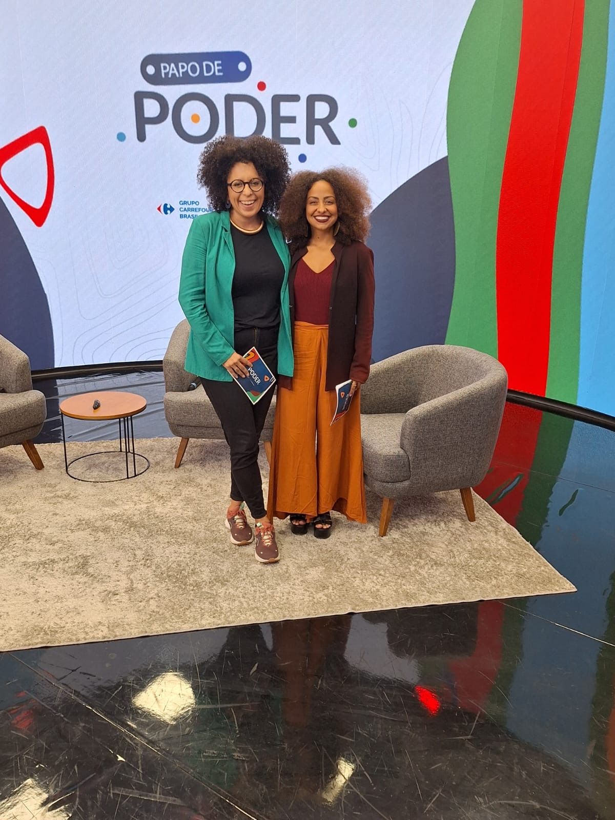 Duas mulheres estão em um cenário. Uma com jaqueta azul-petróleo e calça preta, a outra com roupas marrom-ferrugem.