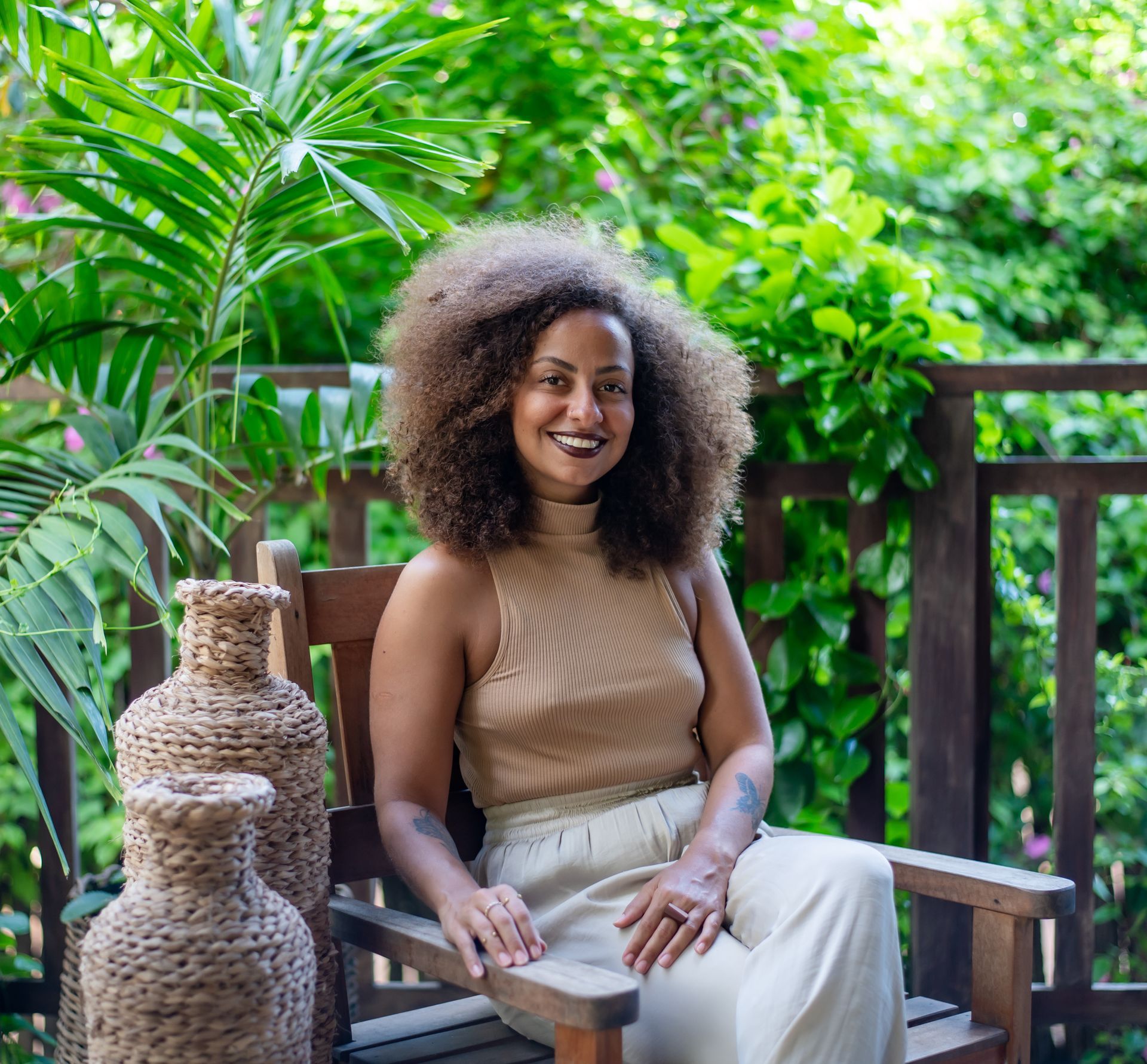 Mulher de cabelos cacheados, vestida com roupa bege, sorri sentada ao ar livre; plantas e vasos de vime estão por perto.