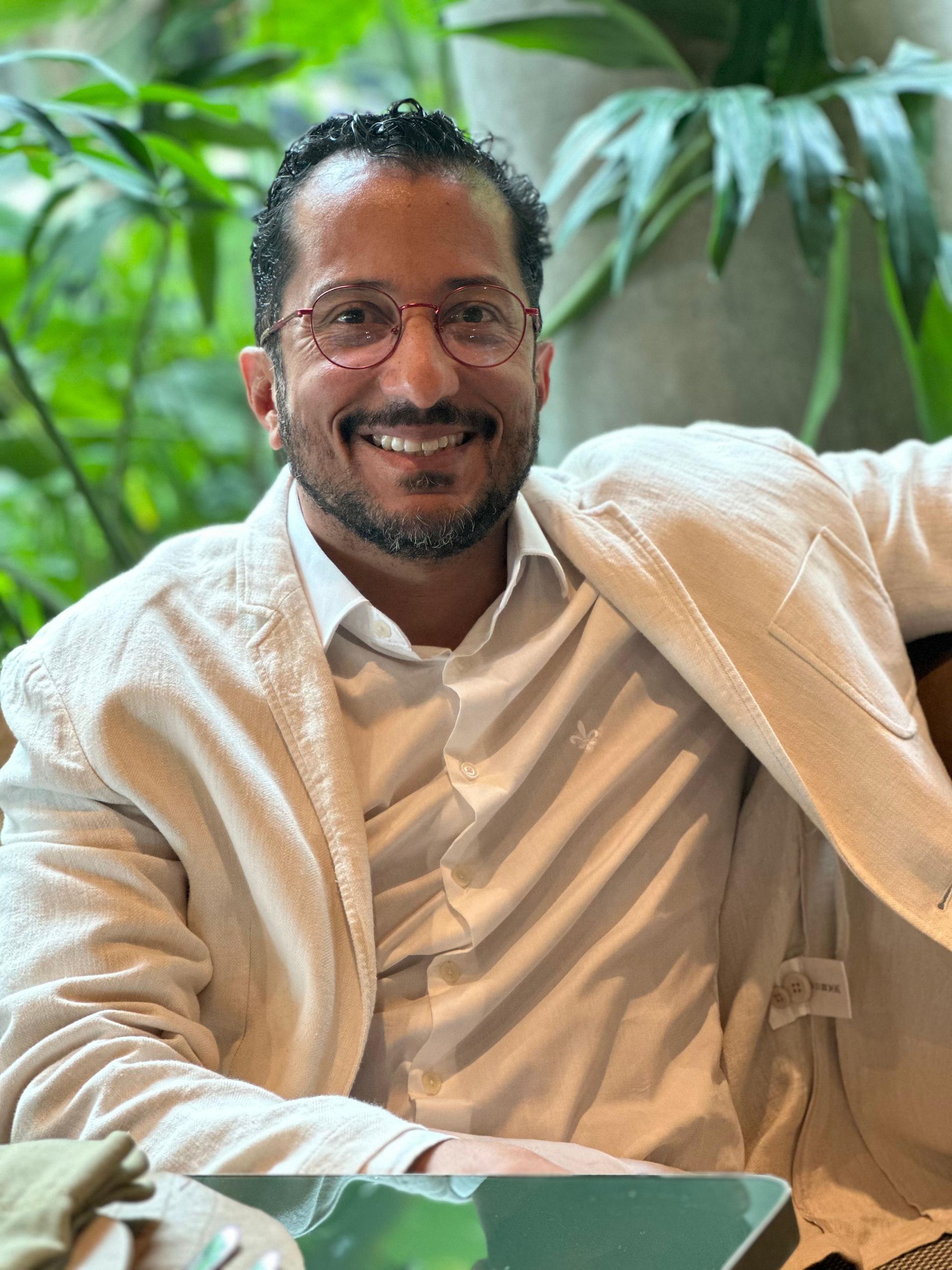 Homem sorridente de óculos e jaqueta leve, fundo de folhagem verde.