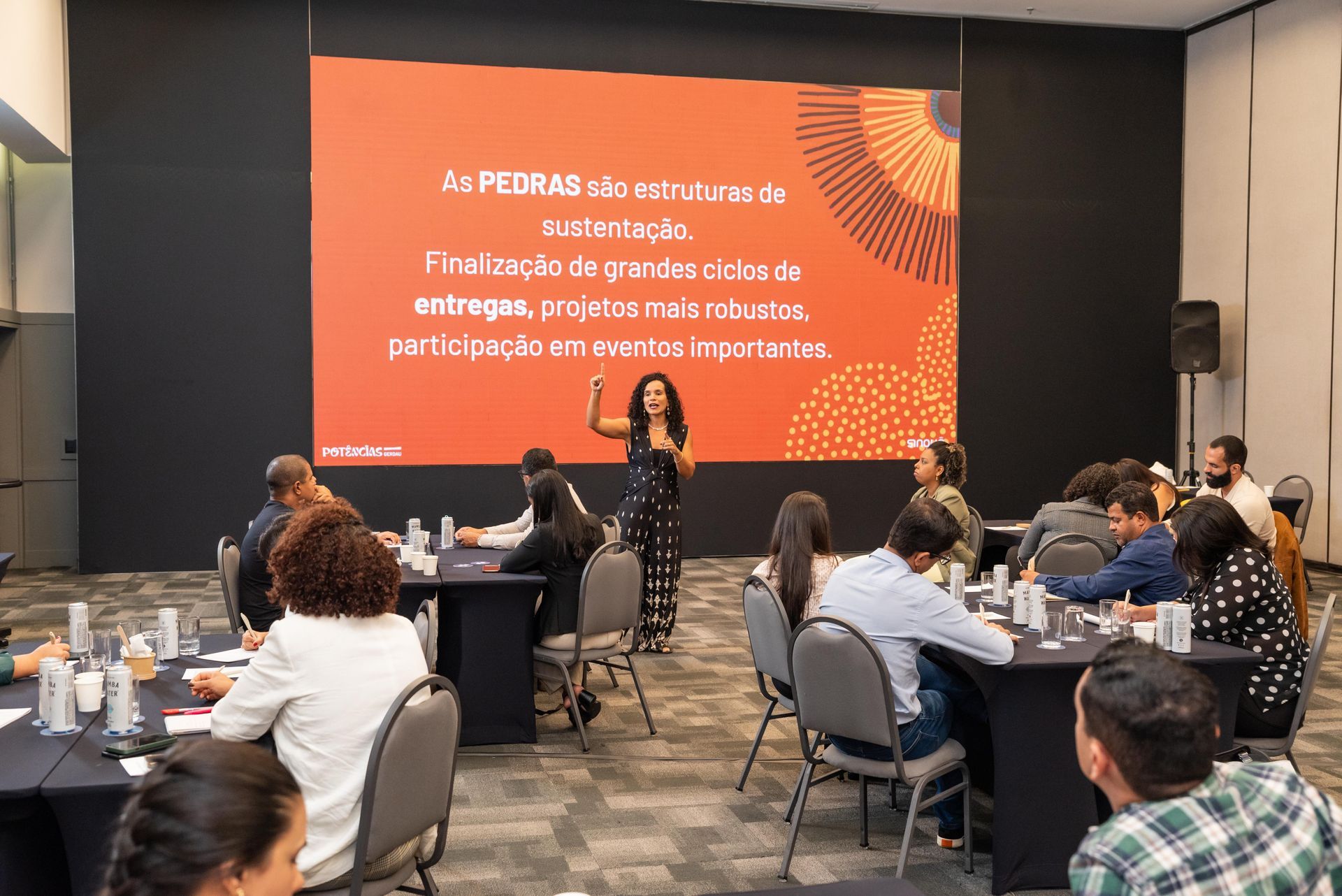 A woman presents to a group at tables. Text on screen: