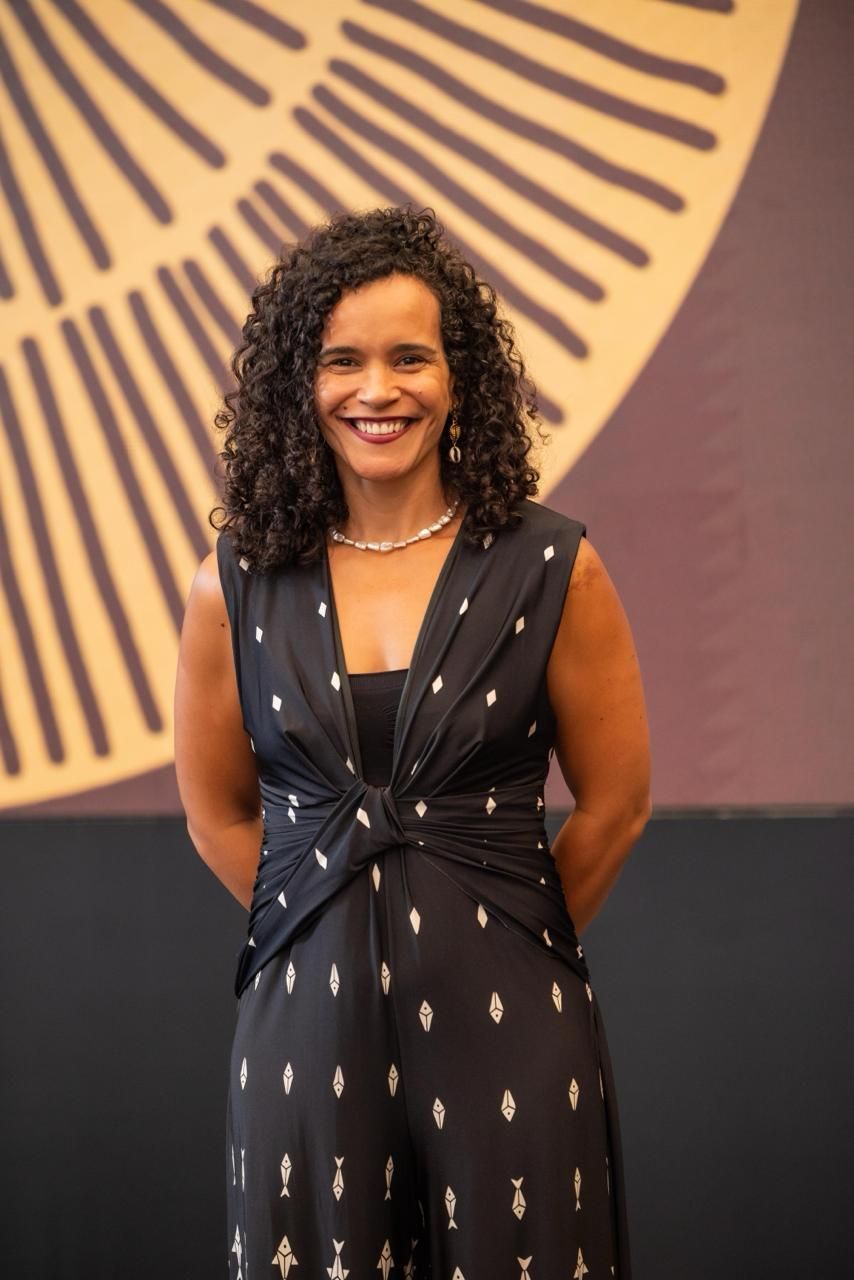 Mulher de macacão preto sem mangas sorri, com os braços atrás das costas, em frente a um fundo marrom abstrato.