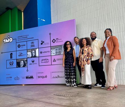 Um grupo de seis pessoas em frente a uma exposição colorida. Elas sorriem. Arquitetura moderna ao fundo.