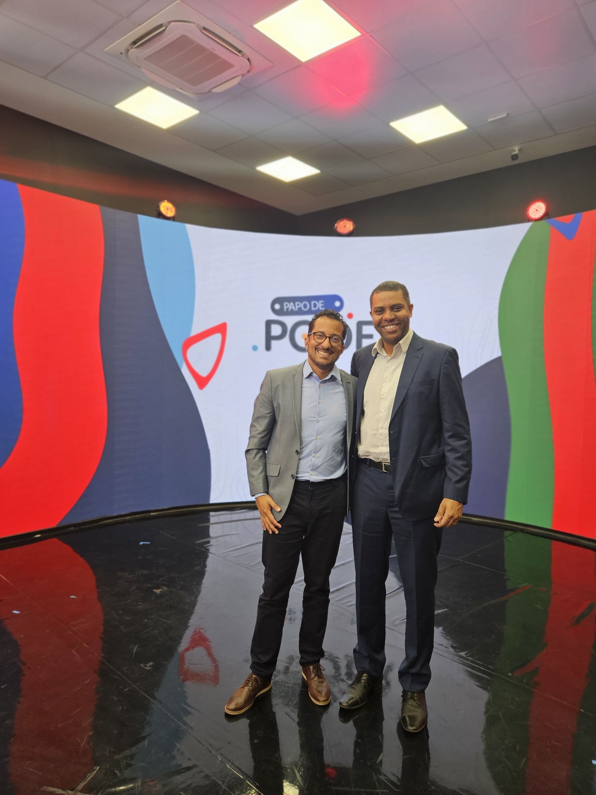 Dois homens de terno estão sorrindo em frente a um aparelho de TV com um fundo curvo, o logotipo