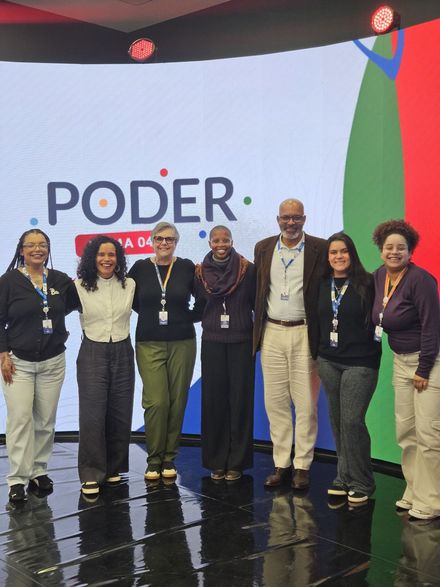Grupo de sete pessoas em pé em um palco com uma placa