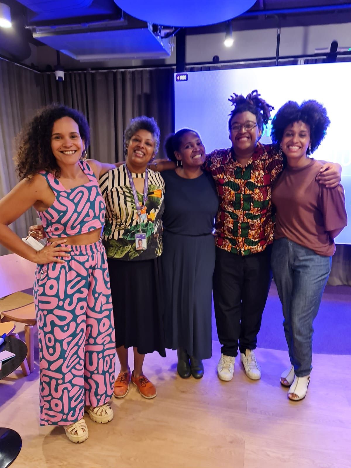 Cinco mulheres, sorrindo, abraçadas, dentro de casa, roupas diferentes, cores quentes, reunião casual.