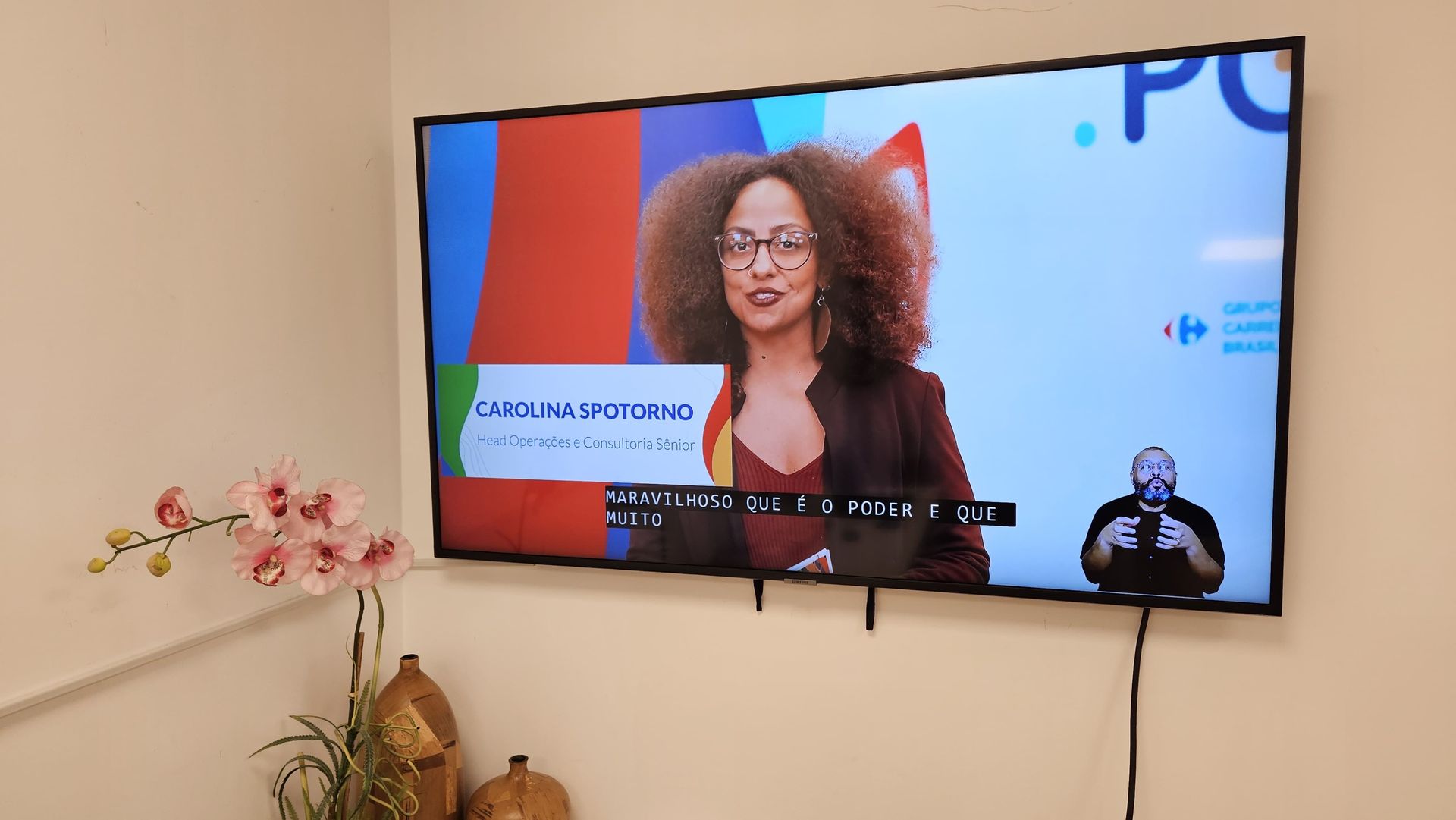 Tela de TV com uma mulher falando, um intérprete de linguagem de sinais e decorações na parede.