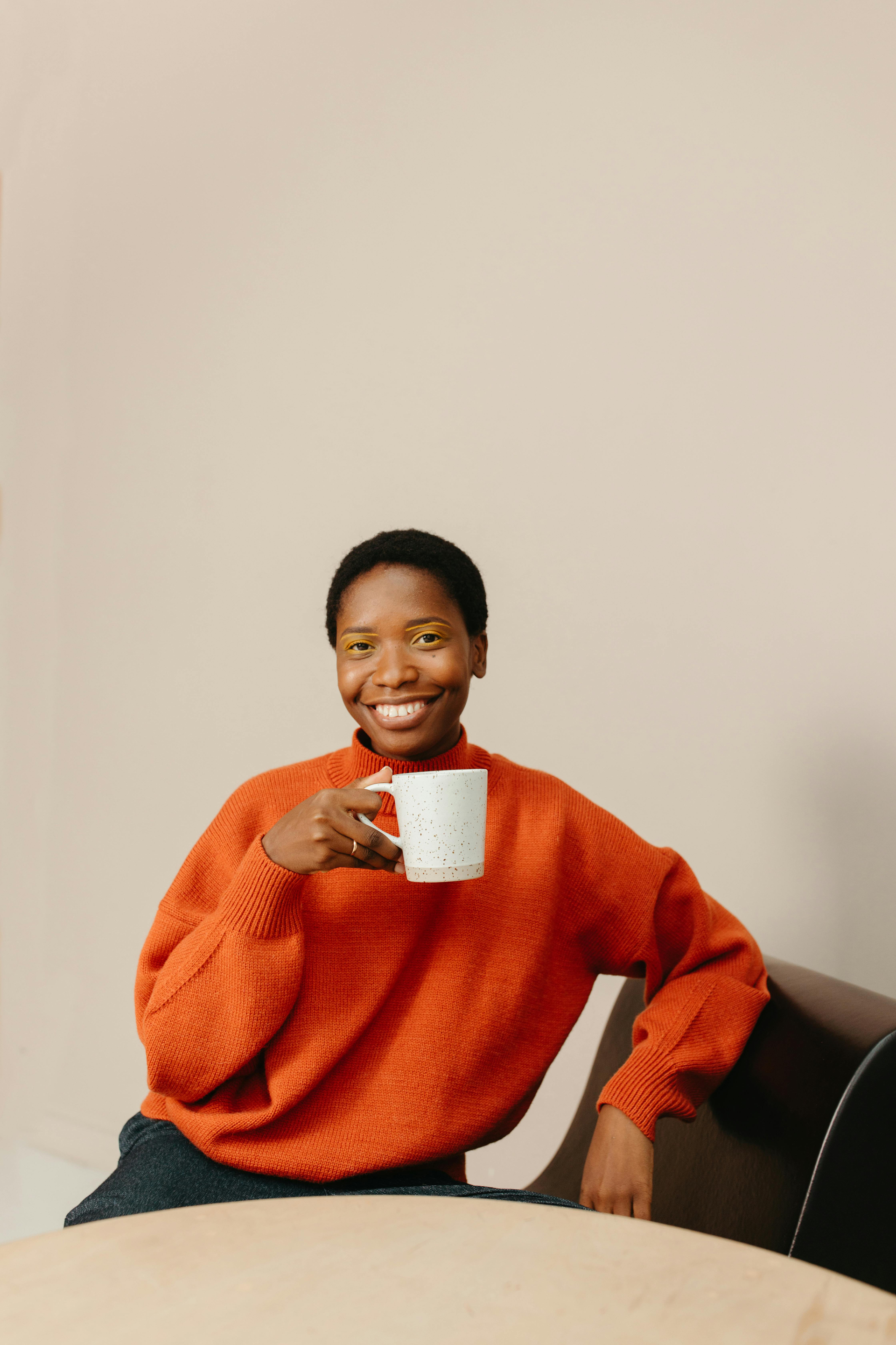 Mulher negra sorridente, de suéter laranja, segurando uma caneca, sentada. Fundo bege.
