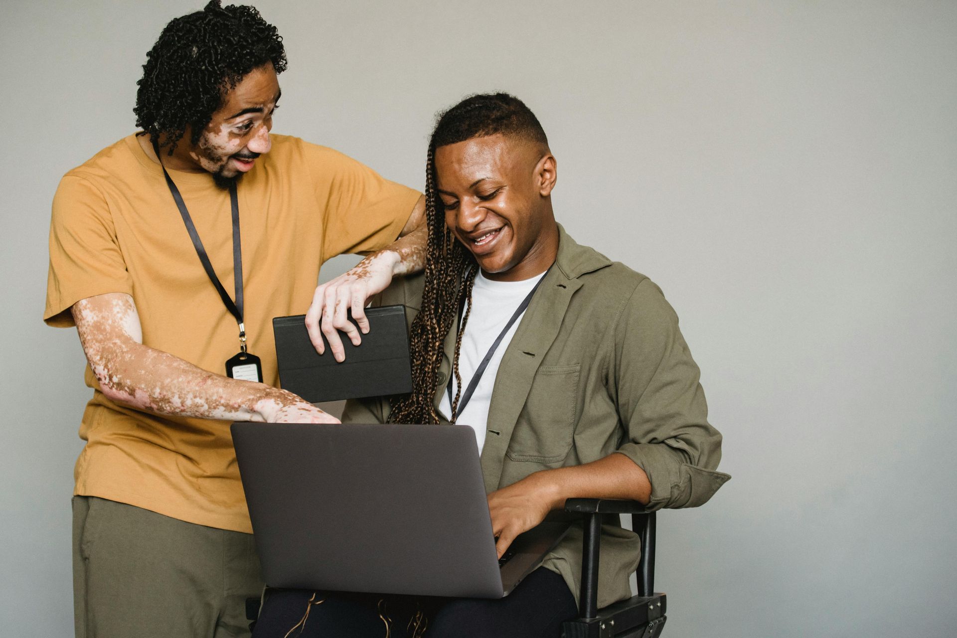 Dois homens olhando para um laptop, um deles em uma cadeira de rodas, sorrindo. Fundo neutro.