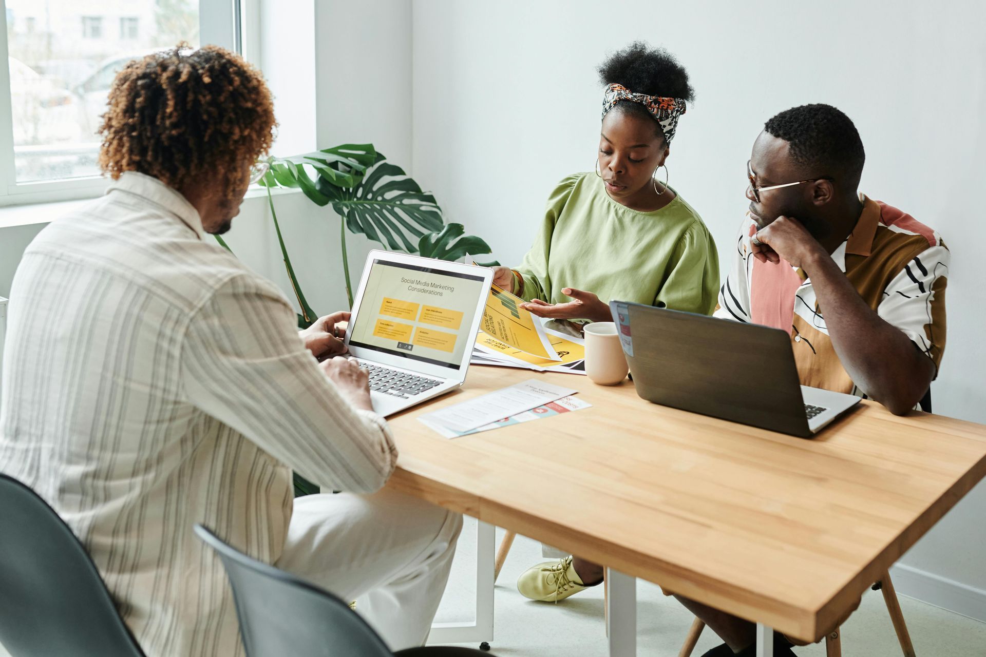 Três pessoas negras sentadas a uma mesa com laptops e papéis. A mulher aponta para um documento, enquanto outros observam.