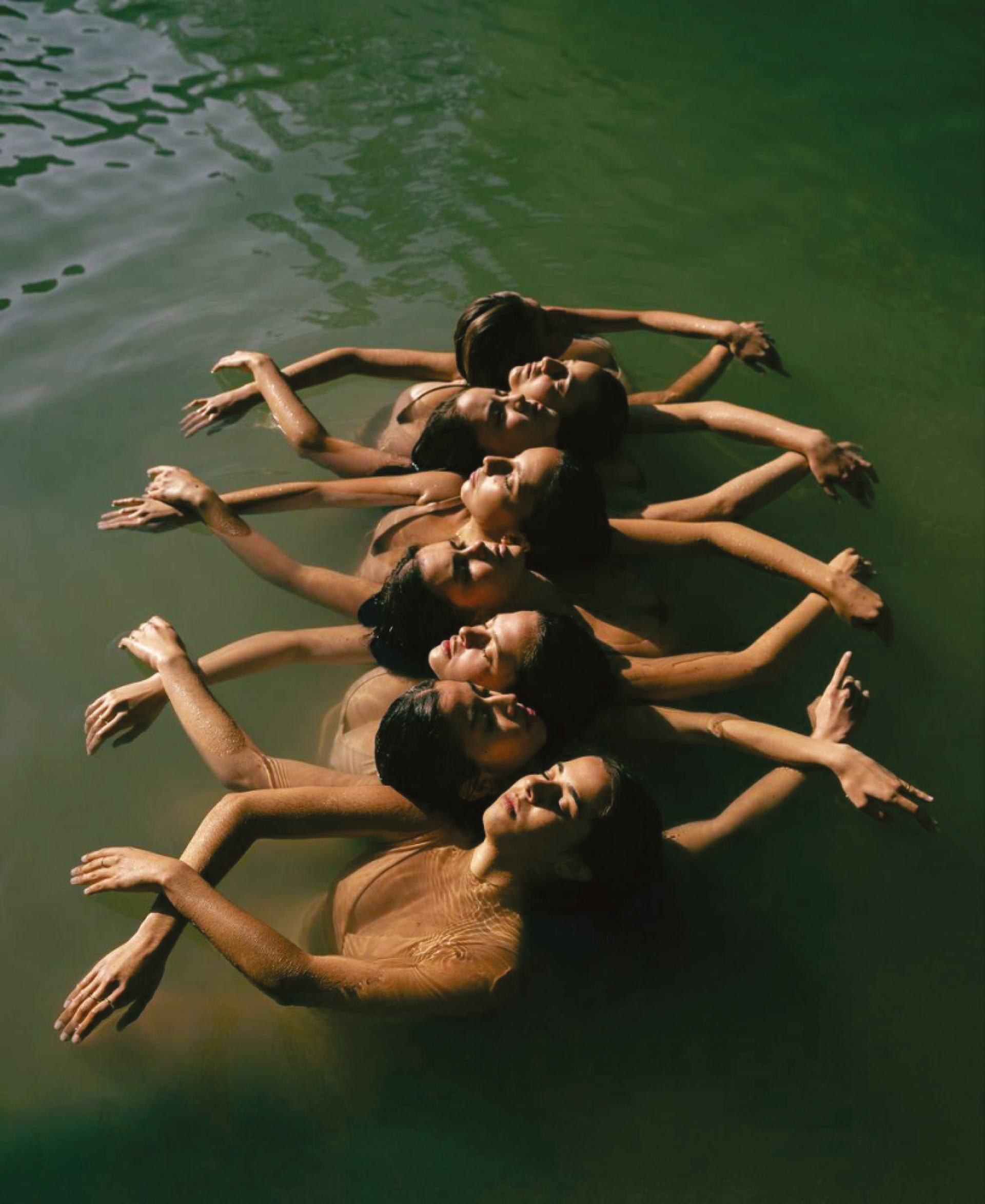 Sete pessoas flutuando em água verde, braços estendidos, rostos inclinados para cima, sorrindo.