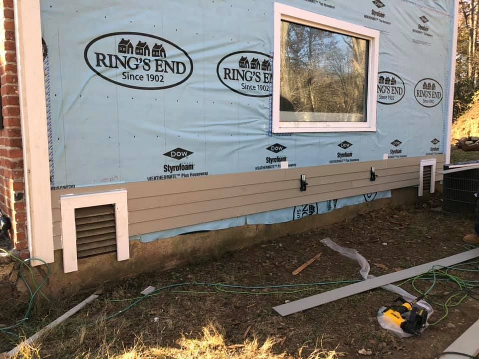 Exterior house siding installation with window and vents. Light brown siding, blue wrap, white trim, and a construction site.