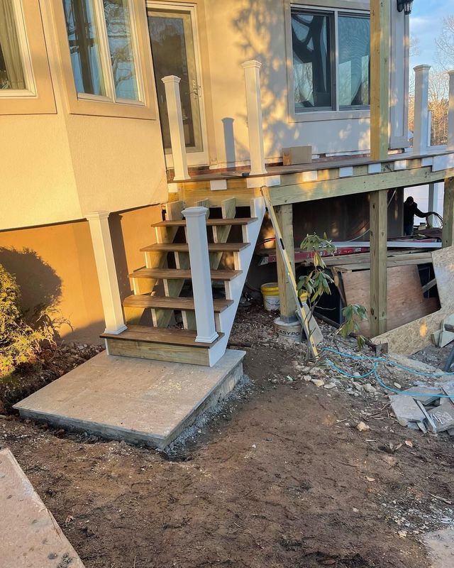 Wooden deck and staircase under construction, built onto the side of a house. Concrete landing at base of stairs.