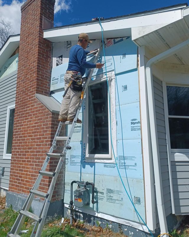 Person on ladder installing siding on house exterior, blue insulation visible.