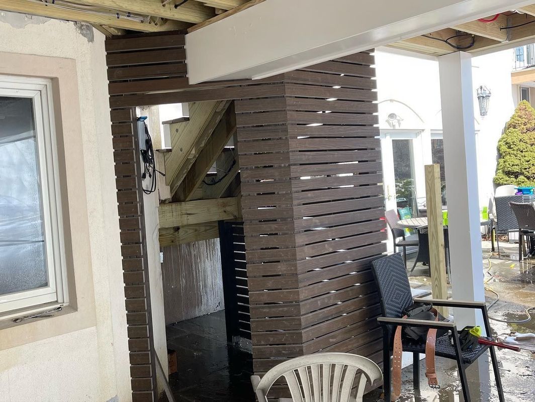 Brown slatted exterior wall with a door, leads to a dark stairwell. A deck and patio are visible.