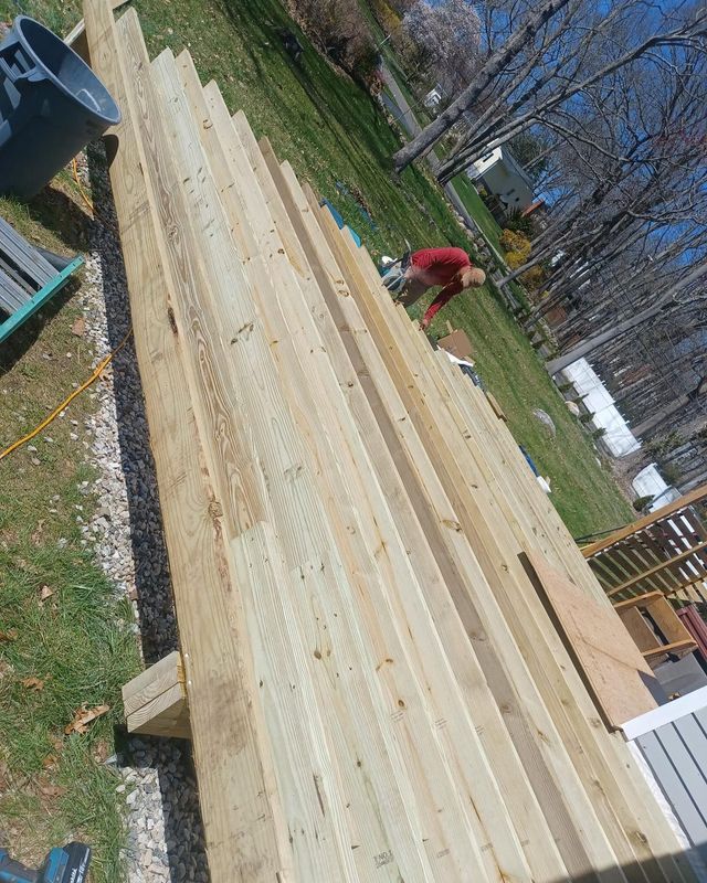 Wooden planks laid out on grass with a person working in the background, near a partially constructed deck.