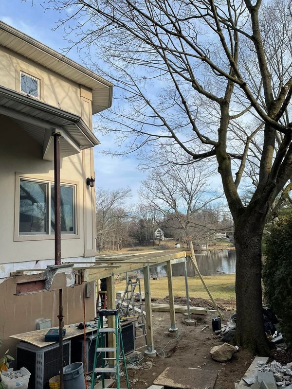 Deck construction near a house and water, with trees and cloudy sky.