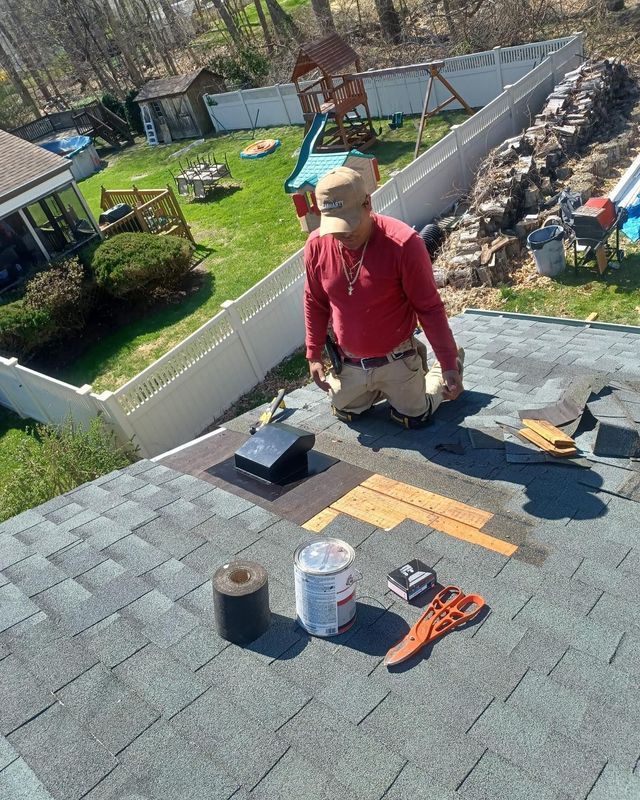 A roofer on a roof repairs shingles. Tools, sealant, and a vent are visible, with a backyard in the background.