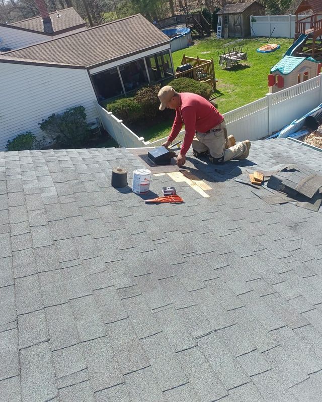 A person kneeling on a gray shingle roof repairs shingles with tools and materials on a sunny day.