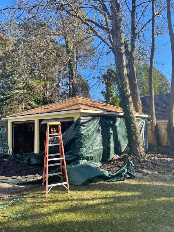 Garage under construction, covered in green tarp. A-frame ladder sits in front. Trees surround the structure.