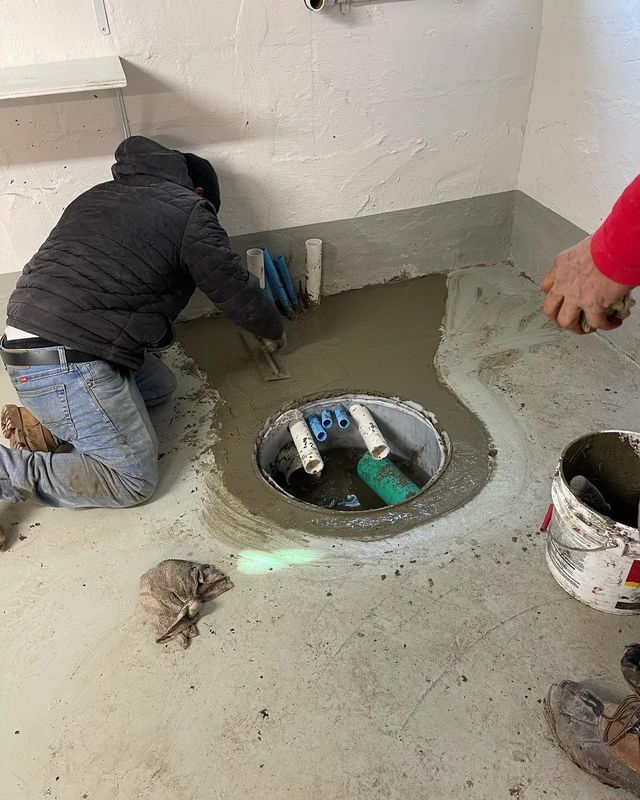 Person kneeling, smoothing concrete around a floor drain. Interior of a utility room with white walls and a gray floor.