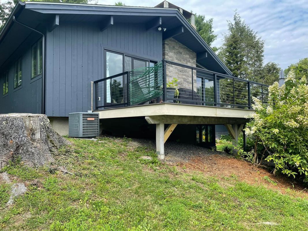 Gray house with a deck supported by wooden posts. Grassy hill in front.