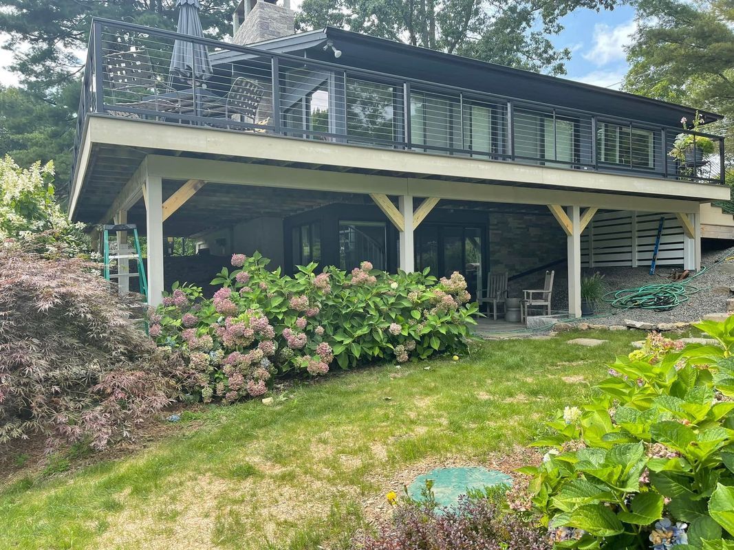 Two-story house with a wooden deck and stone foundation, surrounded by greenery.
