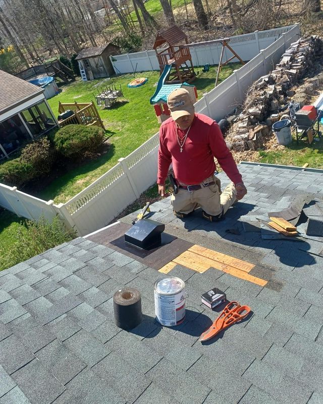 Roofer kneeling on a shingle roof, working with tools. Tools and materials laid out. Sunny backyard visible.