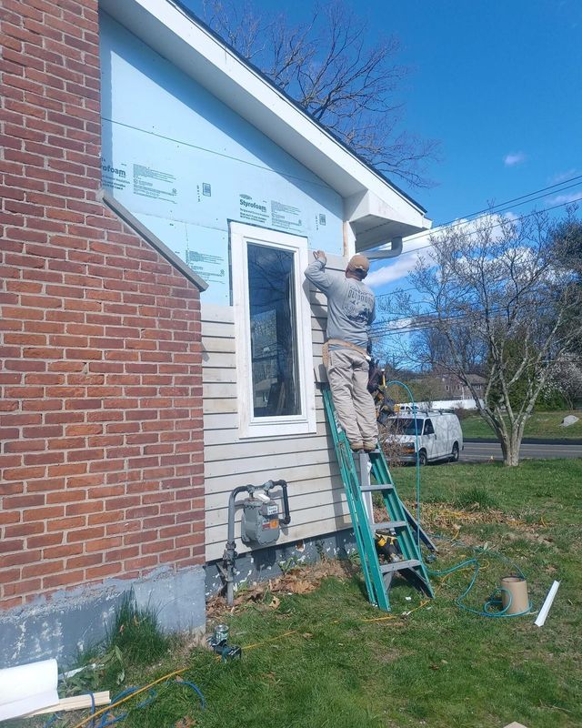 Man on ladder installing siding around a window on a house. Blue sky, brick chimney visible.