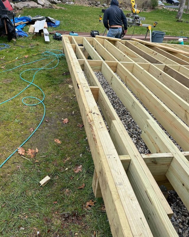 Wooden deck under construction; gravel between beams. A worker screws boards.