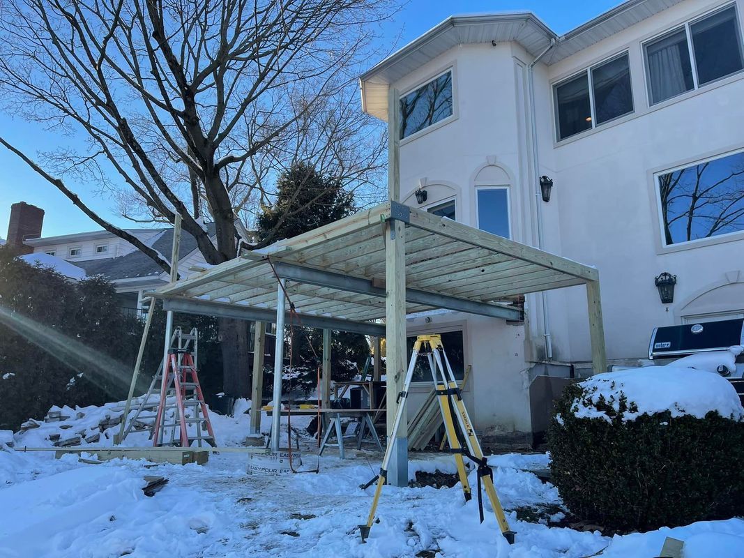 A covered patio under construction in a snowy yard next to a two-story white house.