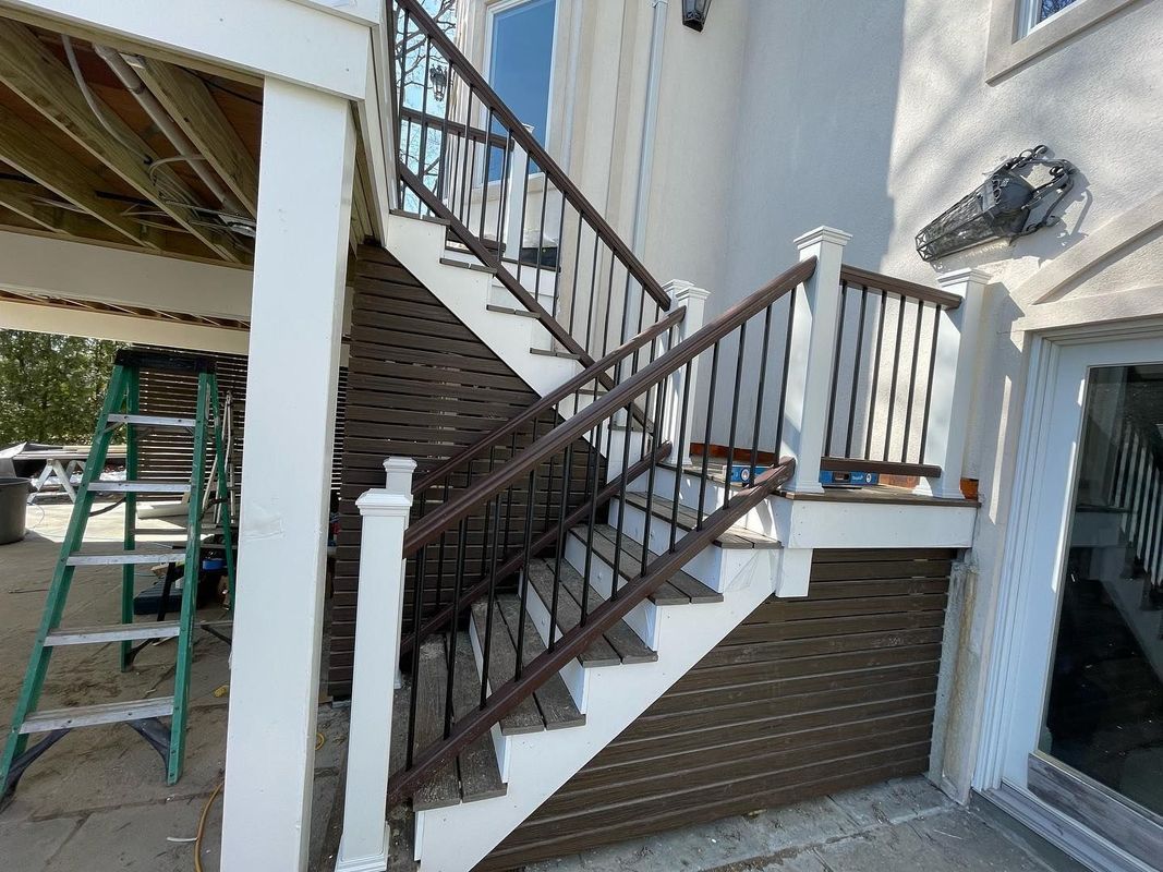 Brown deck stairs with black railings against a white building. A ladder sits to the left.