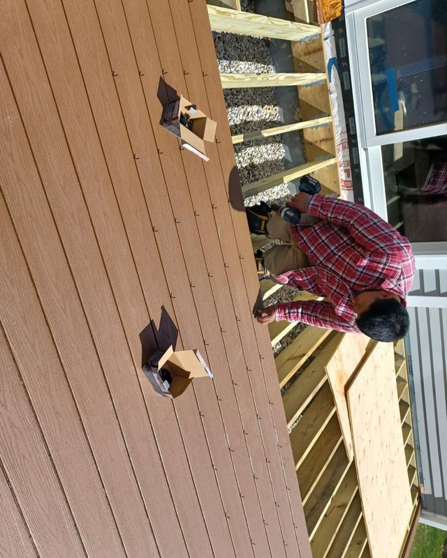 Person in plaid shirt installing deck boards on a wood frame deck. Brown boards, bright sun.