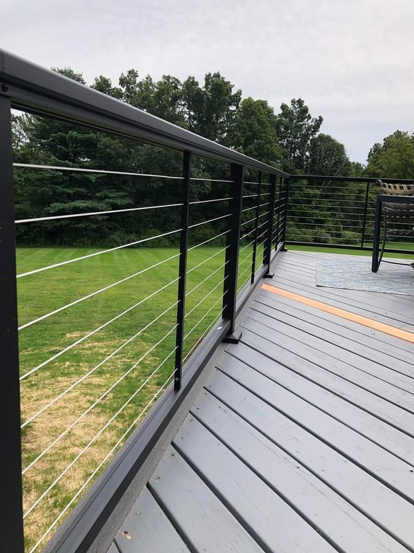 Black metal deck railing with horizontal steel cables on a grey wooden deck overlooking a green lawn.