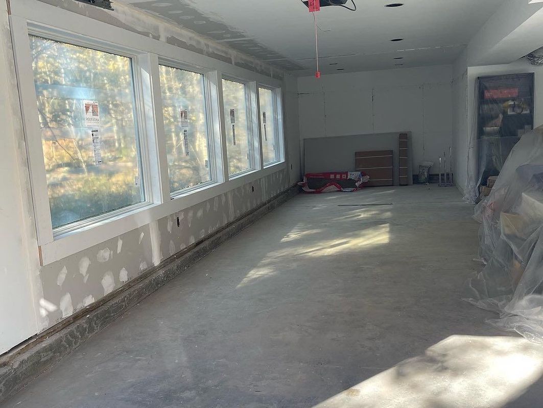 Interior room under construction with long row of windows and concrete floor.