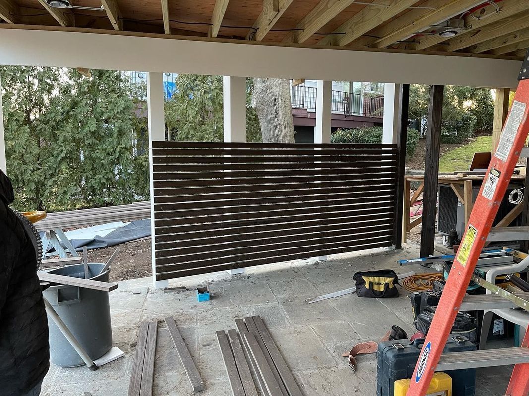 Construction site under a deck with a horizontal slat privacy screen; brown, white, and gray colors.