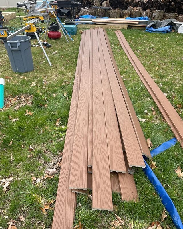 Pile of brown composite deck boards on green grass. Construction tools visible in the background.