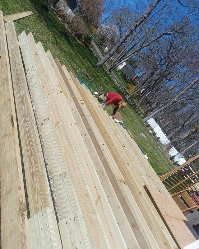 Person working on a deck, surrounded by wooden planks, on grass. The sky is visible.