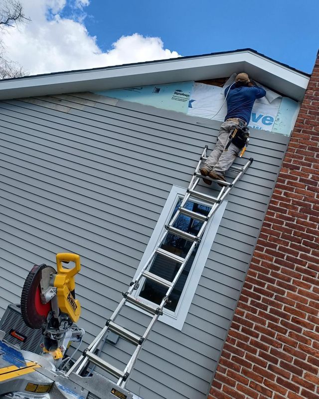 Person on ladder working on siding of a house, using a saw with blue sky.