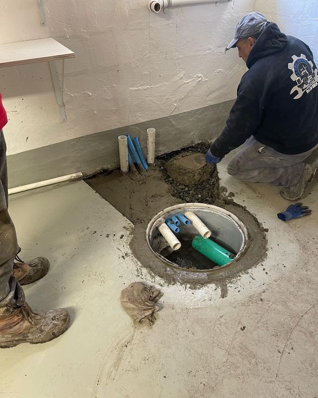 Plumber working on a sump pump installation in a concrete basement. Grey and blue pipes, cement, man in a hoodie.