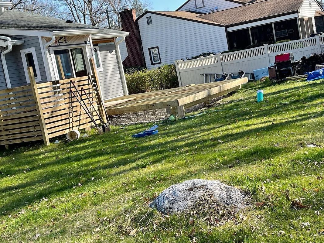 A partially constructed wooden deck in a grassy yard, with a small house and other houses in the background.
