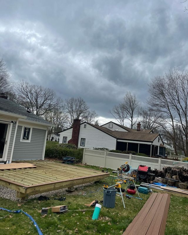 Backyard deck under construction; cloudy sky. Houses in the background. Construction tools and materials scattered on the grass.