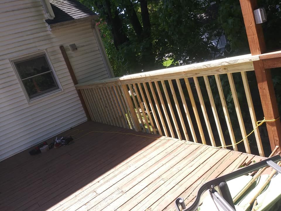 Wooden deck with railing, attached to a house. Sunlight shines on the deck and surrounding trees.