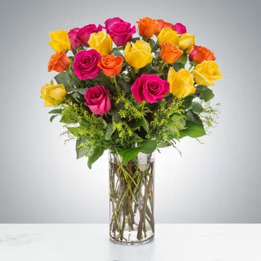 A tall clear glass vase holding a vibrant bouquet of yellow, orange, and magenta roses with lush green foliage.