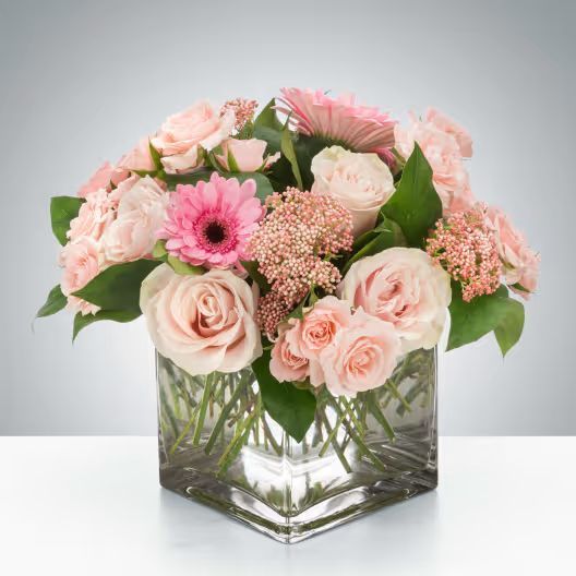 A bouquet of pink roses, gerbera daisies, and filler flowers arranged in a square glass vase against a gray background.