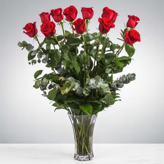 A bouquet of red roses with green eucalyptus leaves in a clear glass vase against a plain, light background.