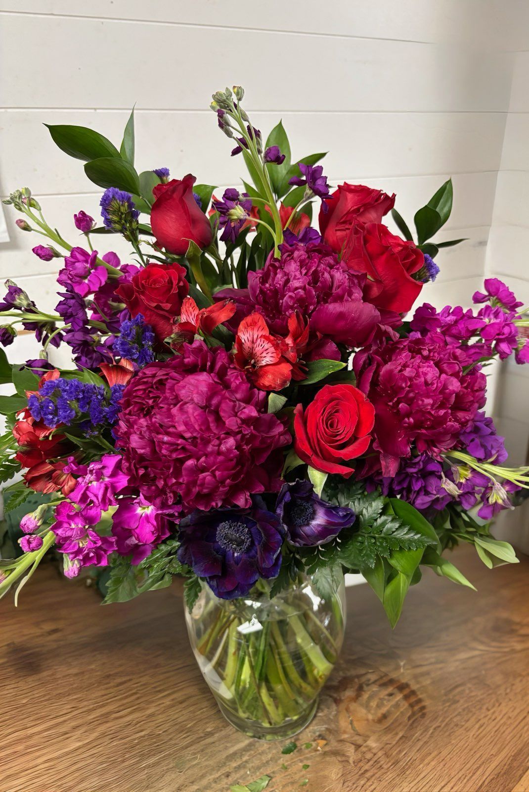 A vibrant bouquet of deep red roses, magenta peonies, and purple stock flowers in a clear glass vase on a wooden table.