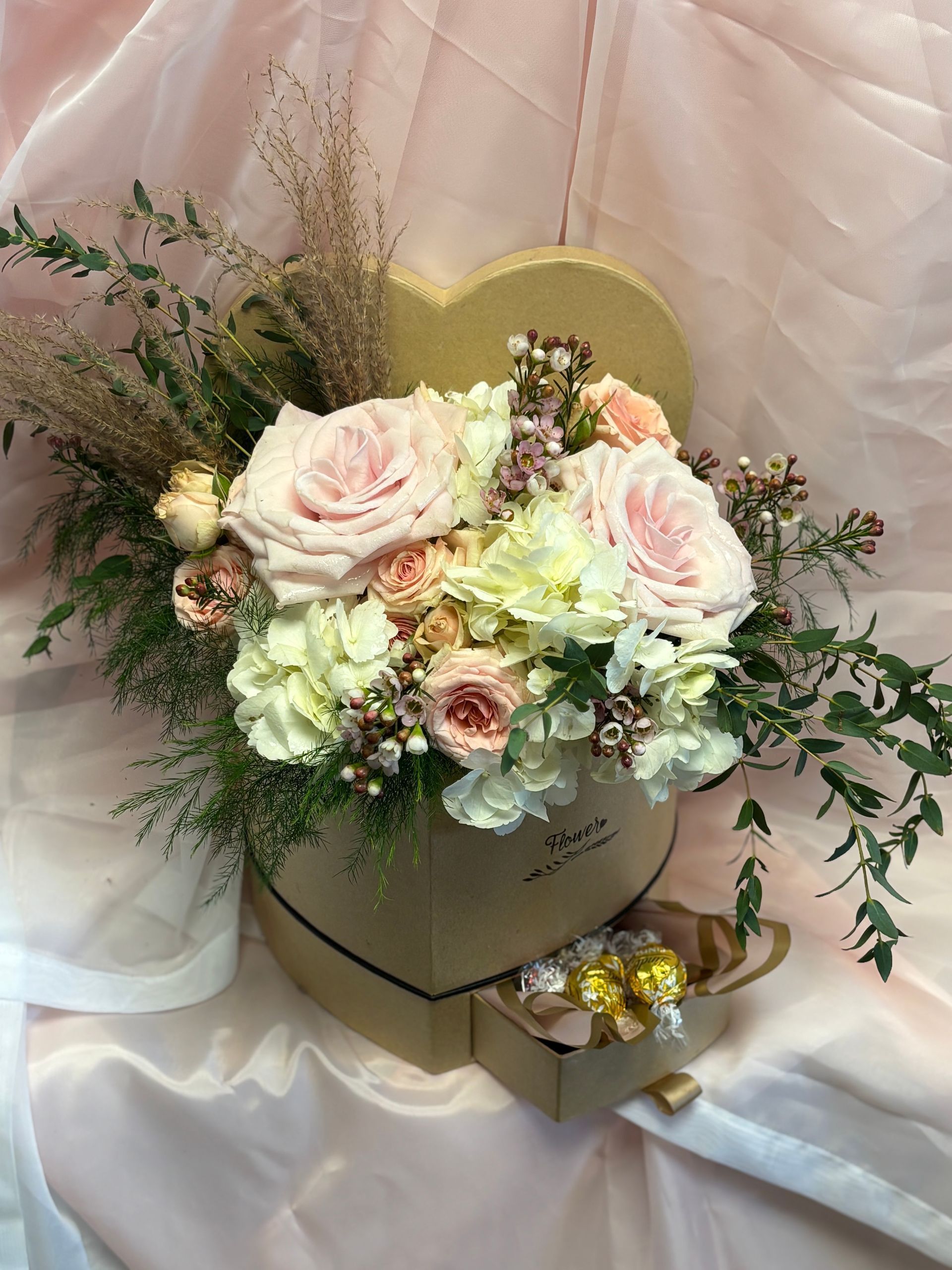 A heart-shaped gift box filled with blush roses, white hydrangeas, and greenery, with a small drawer containing candy.