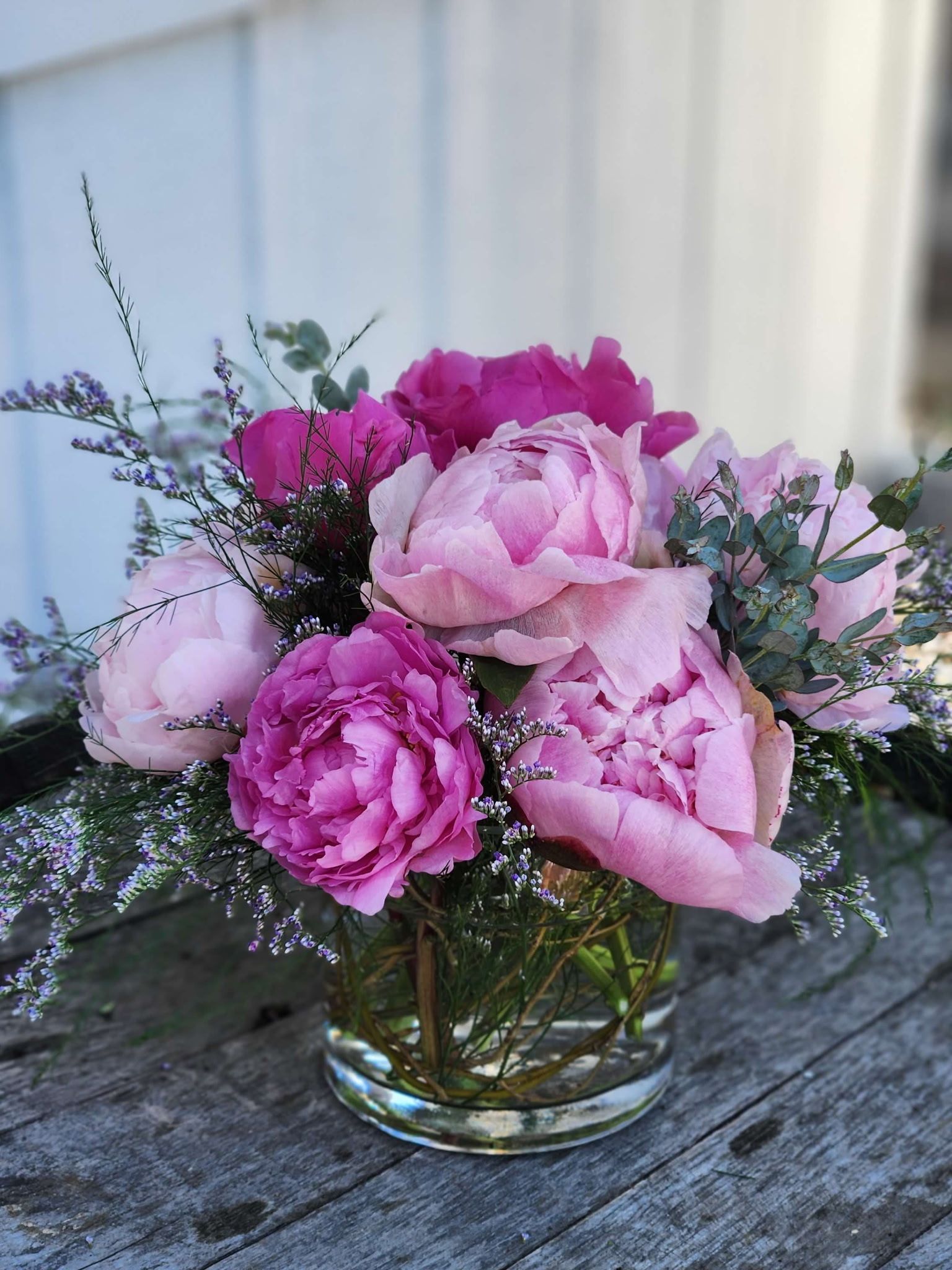 A vibrant bouquet of pink and magenta peonies with green foliage in a glass vase on a rustic wooden surface.