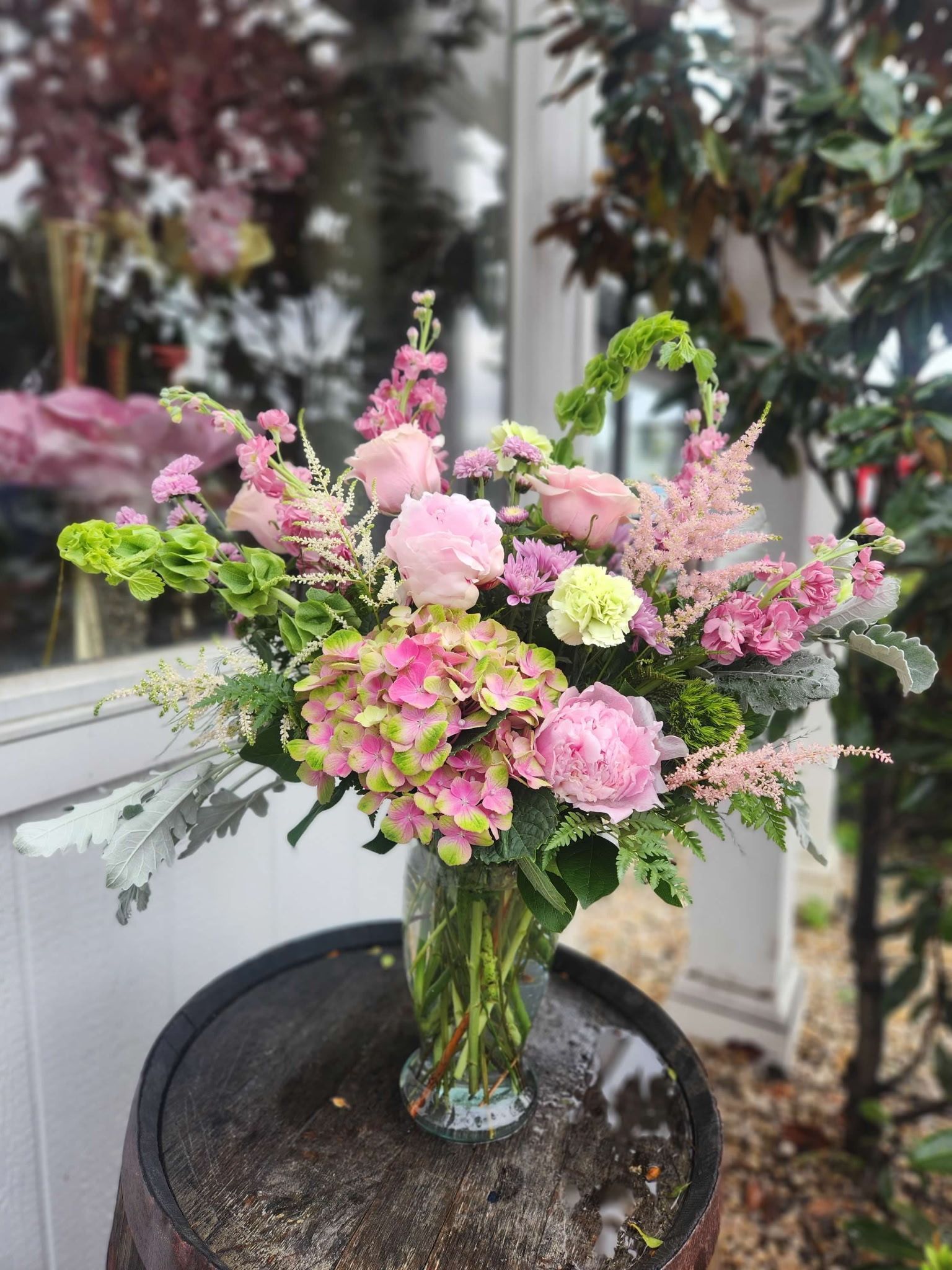 A lush, vibrant floral arrangement in a clear glass vase sits atop a dark wooden barrel outdoors.