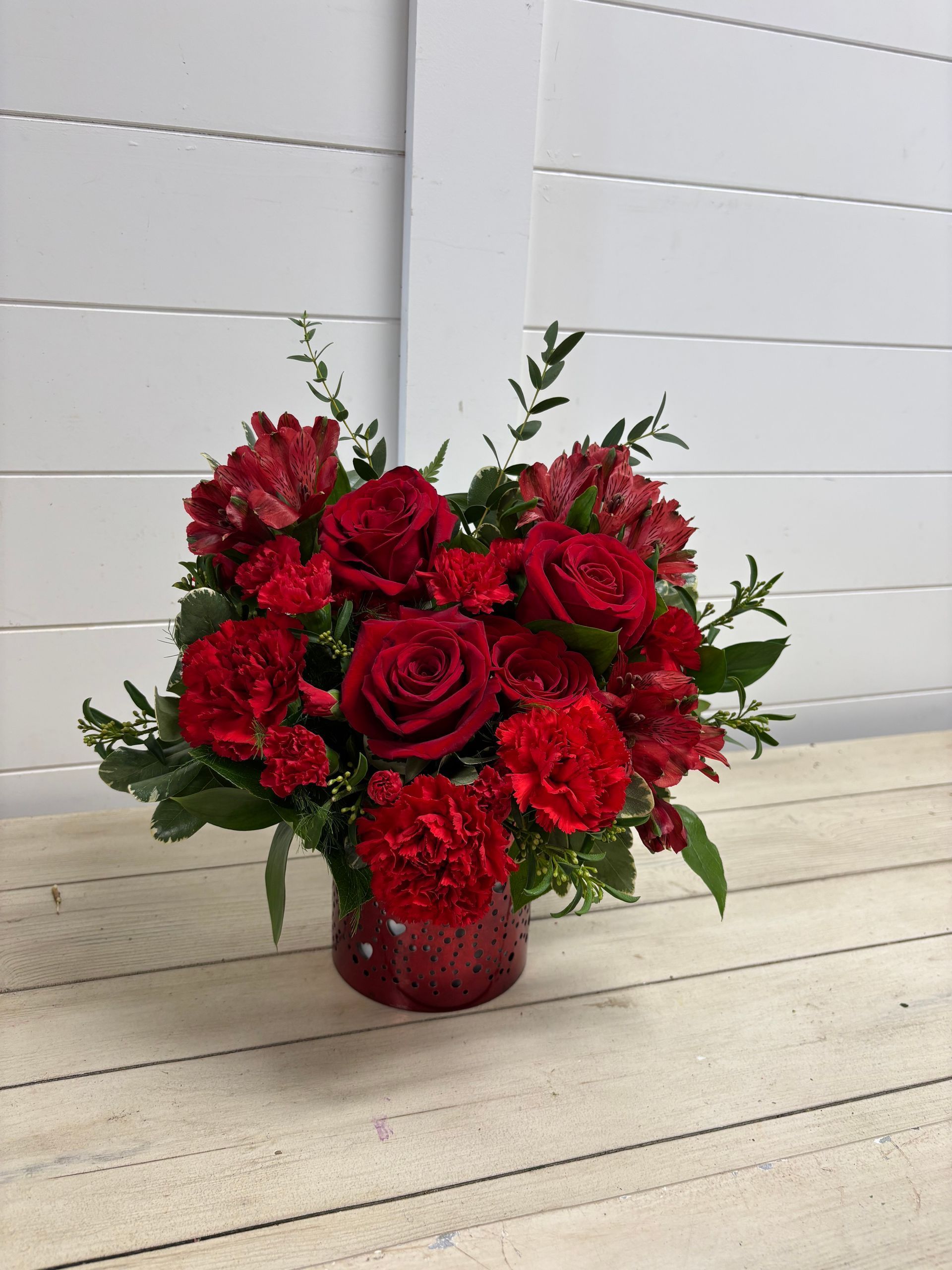 A lush arrangement of red roses, carnations, and greenery in a textured red glass vase on a white wooden surface.